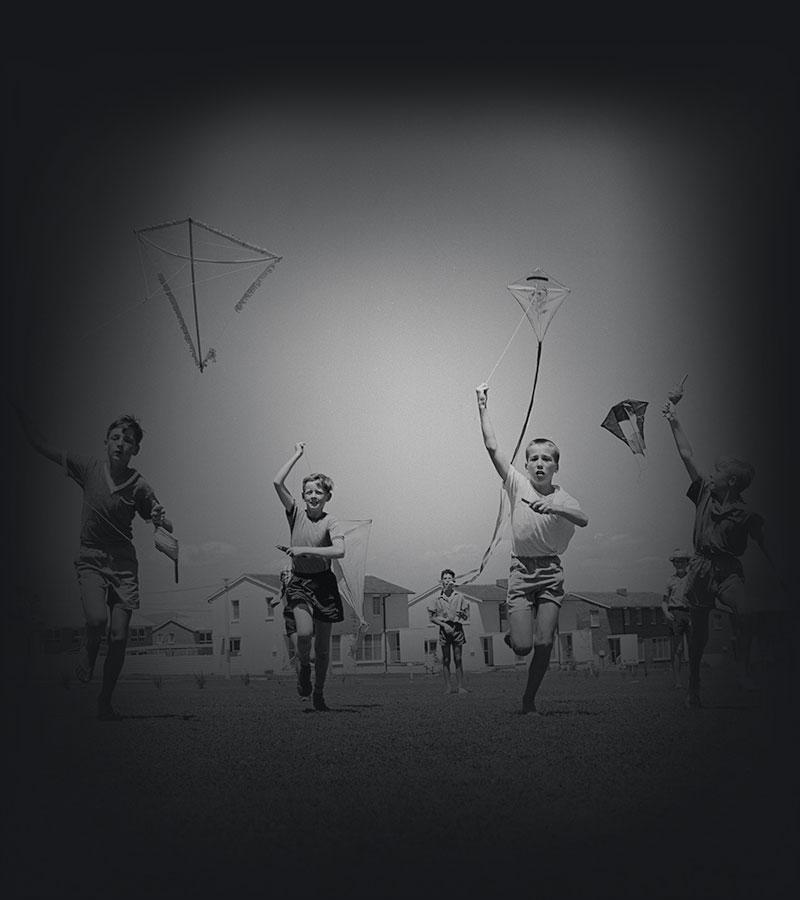 Four children running toward the camera with kites while four children watch from behind.