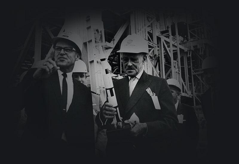 Men in suits wearing hard hats standing before a rocket gantry.