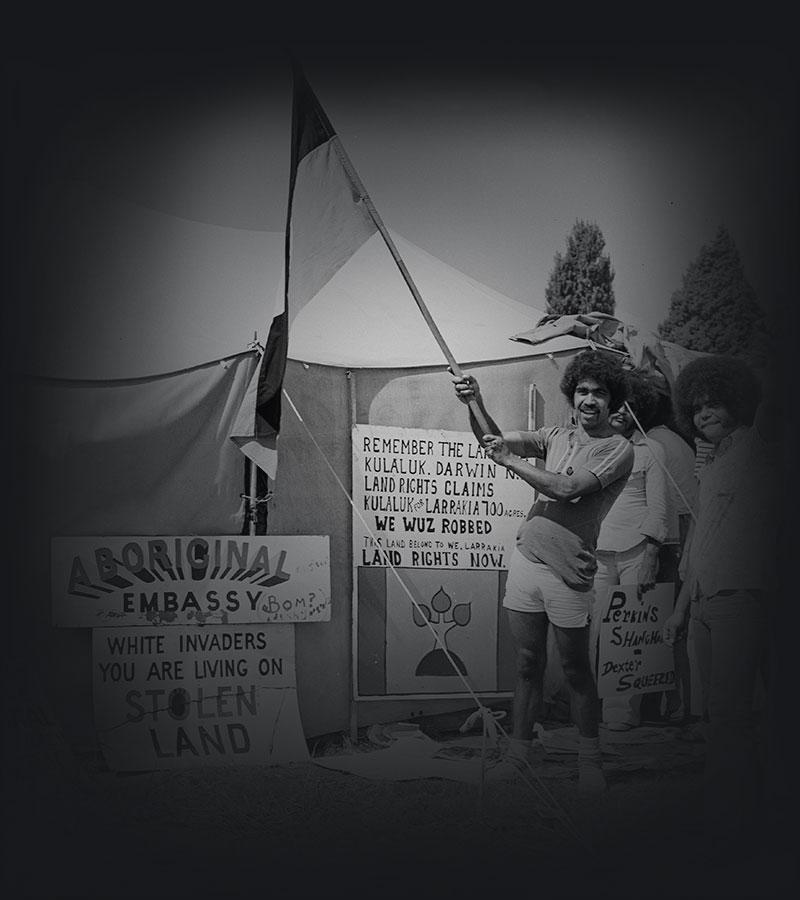 Two men standing outside the Aboriginal Tent Embassy, one holding an Aboriginal flag. 