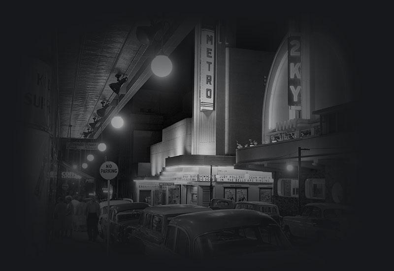 Night street view of pedestrians, parked cars, Metro Theatre and 2KY radio.