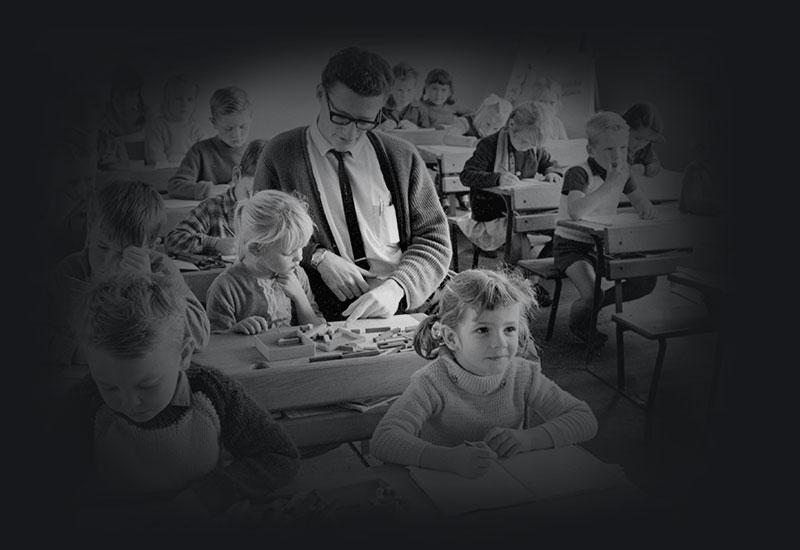 Young children sitting at desks in a classroom while a teacher assists one of the students.