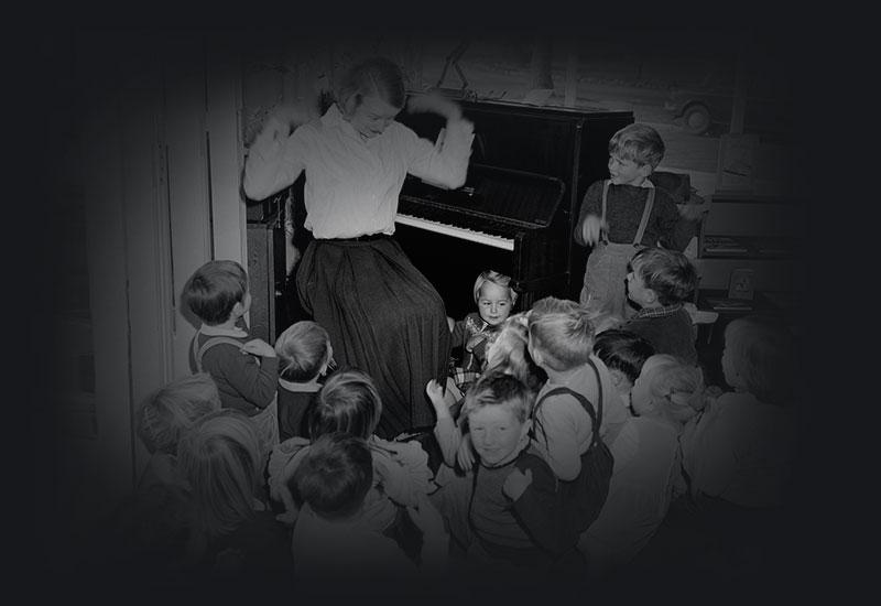 A class of young children gathered around a teacher seated beside a piano shaking her hands.