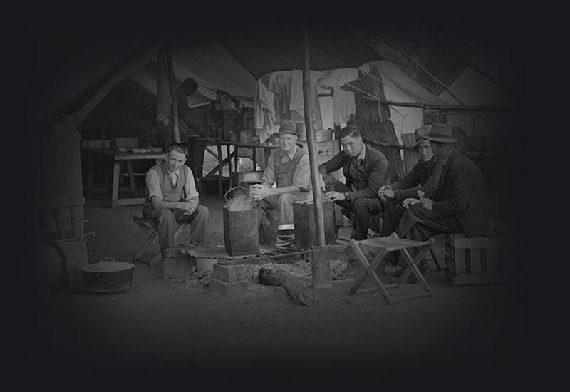 5 men sitting around an open fire heating tin buckets in front of an open mess tent.