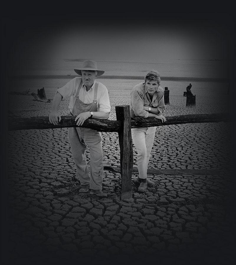 2 farmers leaning against a timber rail fence examining the parched ground of a dried-up dam