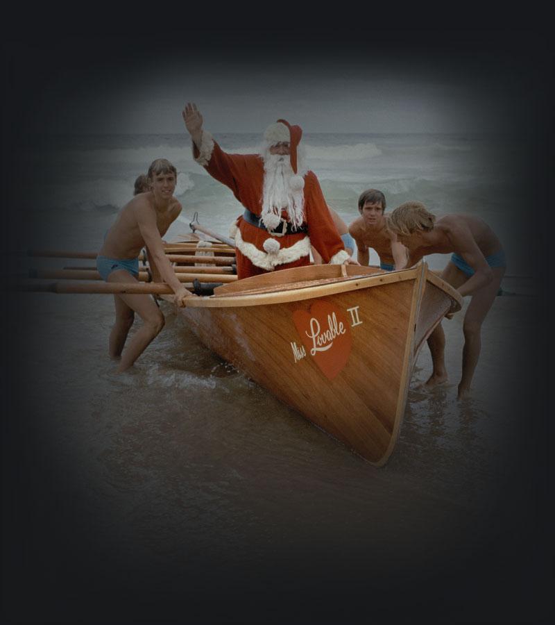 Surf lifesavers push boat carrying man in Santa suit onto sand.