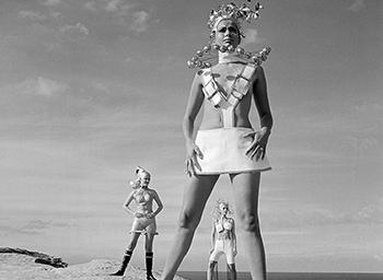 Three women standing on a sandstone cliff above the ocean dressed in futuristic fashion.