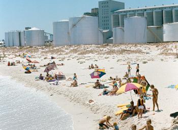 People sun baking on a beach overlooked by steel and concrete silos behind..