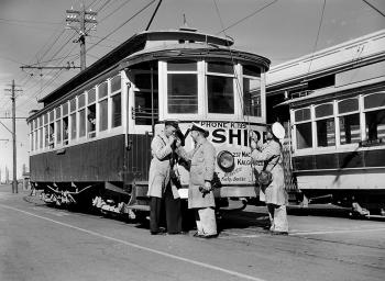 2 tram drivers enjoy a smoko while a colleague adjusts the tram pole and passengers look at the photographer through the tram windows.