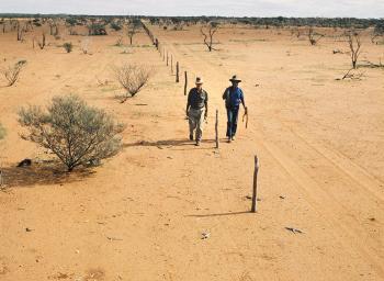 2 people walk a fence line across an arid laandscape of red dirt.