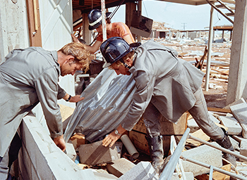 3 men search under corrugated steel and concrete bricks.