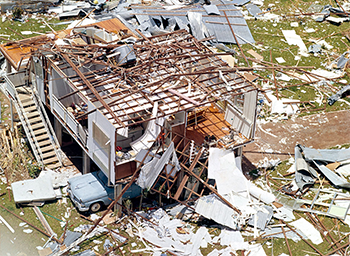 Cars parked beneath 2 destroyed houses surrounded by housing material.