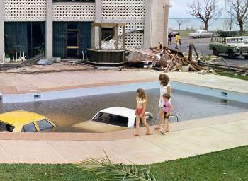 A woman and 2 children walk by 2 submerged cars in a multistorey hotel pool.