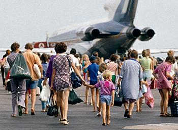 People in colourful clothing walk toward an aircraft with letter 'T' on the tail.