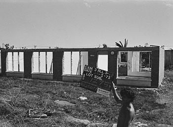 A man holds a land allotment sign infront of a damaged house.