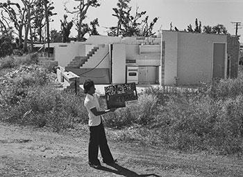 A man holds a land allotment sign infront of a damaged house.