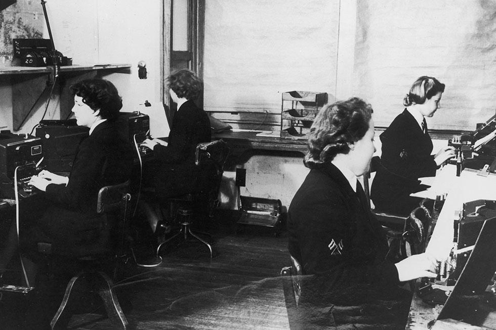 Four women in uniform seated at desks operating code machines.