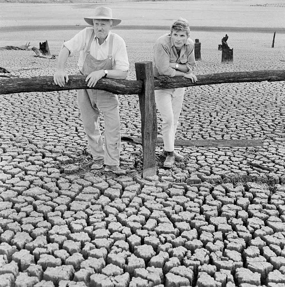 Two men leaning on a timber post and rail fence looking at the dry ground cracked into large blocks.