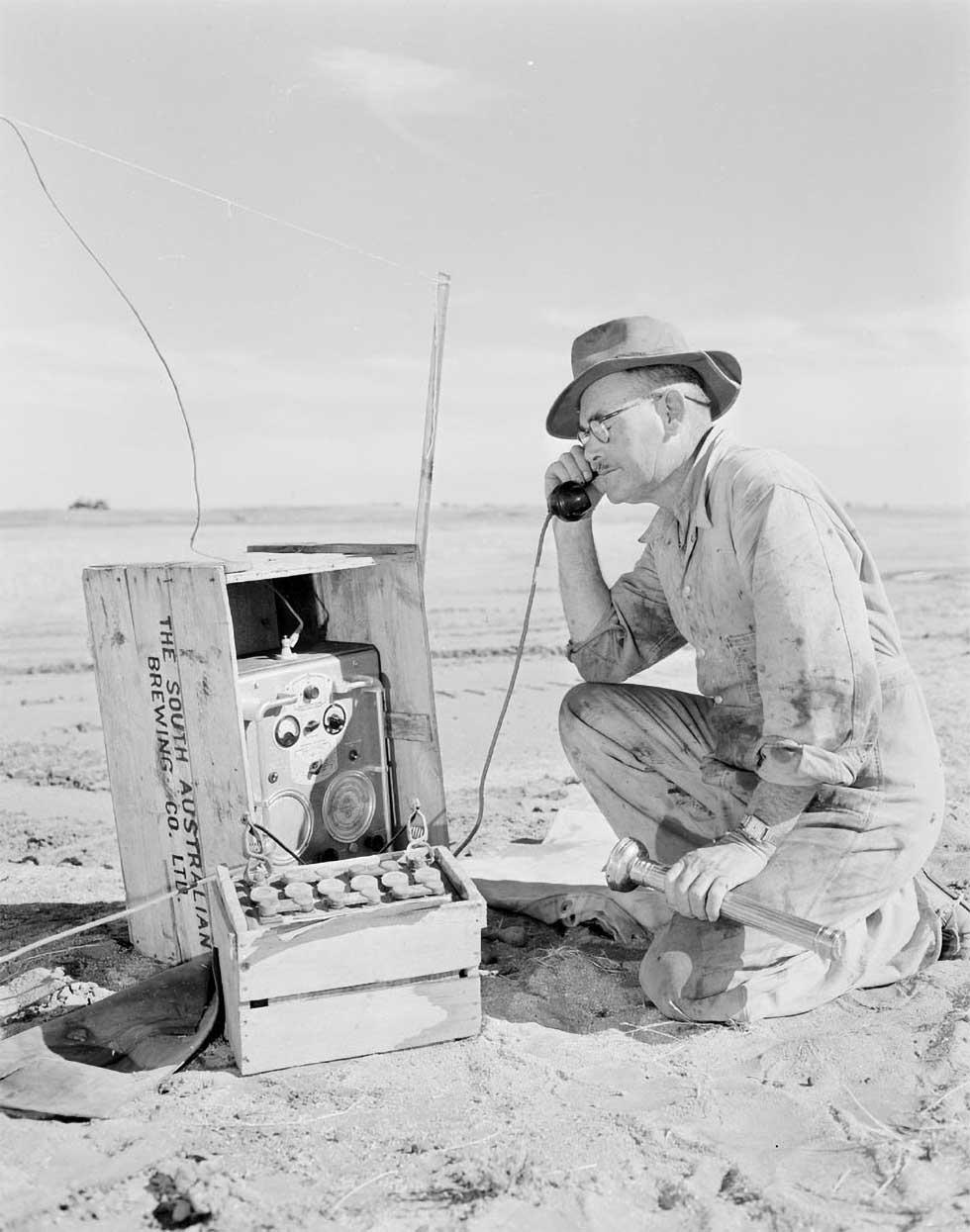 George Alder in a flat, dry and dusty paddock kneeling beside the first portable radio telephone in the outback.