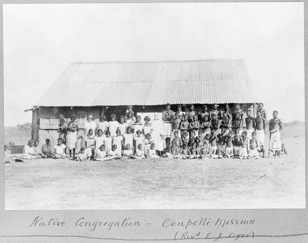 This black-and-white photograph shows two groups of First Australians, posing in front of a hut with a tin roof. Women and girls in white dresses stand on one side of the photo and shirtless men and boys in long white shorts on the other. In the middle of the photo is a clergyman in a white surplice. His wife stands to the left, among the girls and women. The photo is inscribed ‘Native Congregation — Oenpelli Mission (Rev’d E. J. Dyer)’.