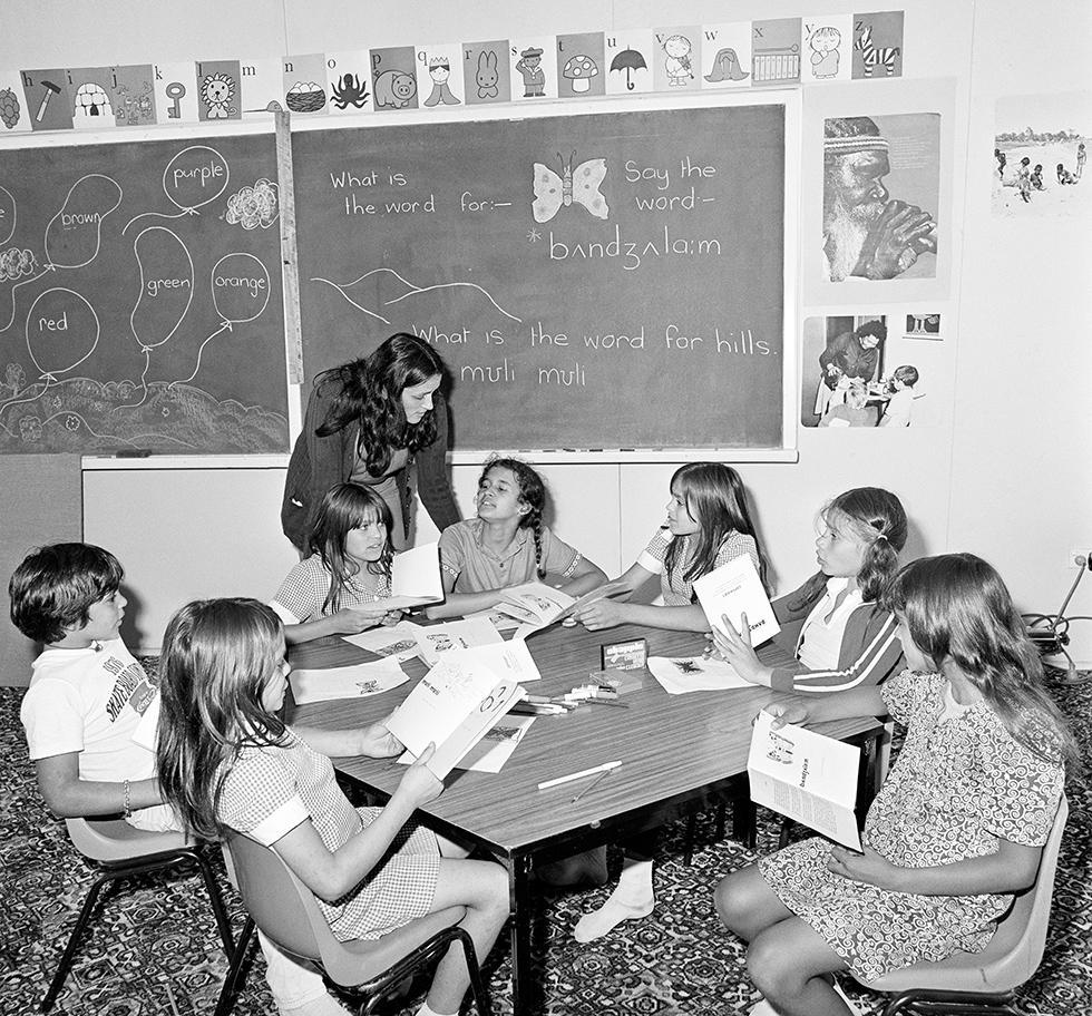 7 school children seated around a table listening to their teacher. A blackboard features drawings of hills and a butterfly.