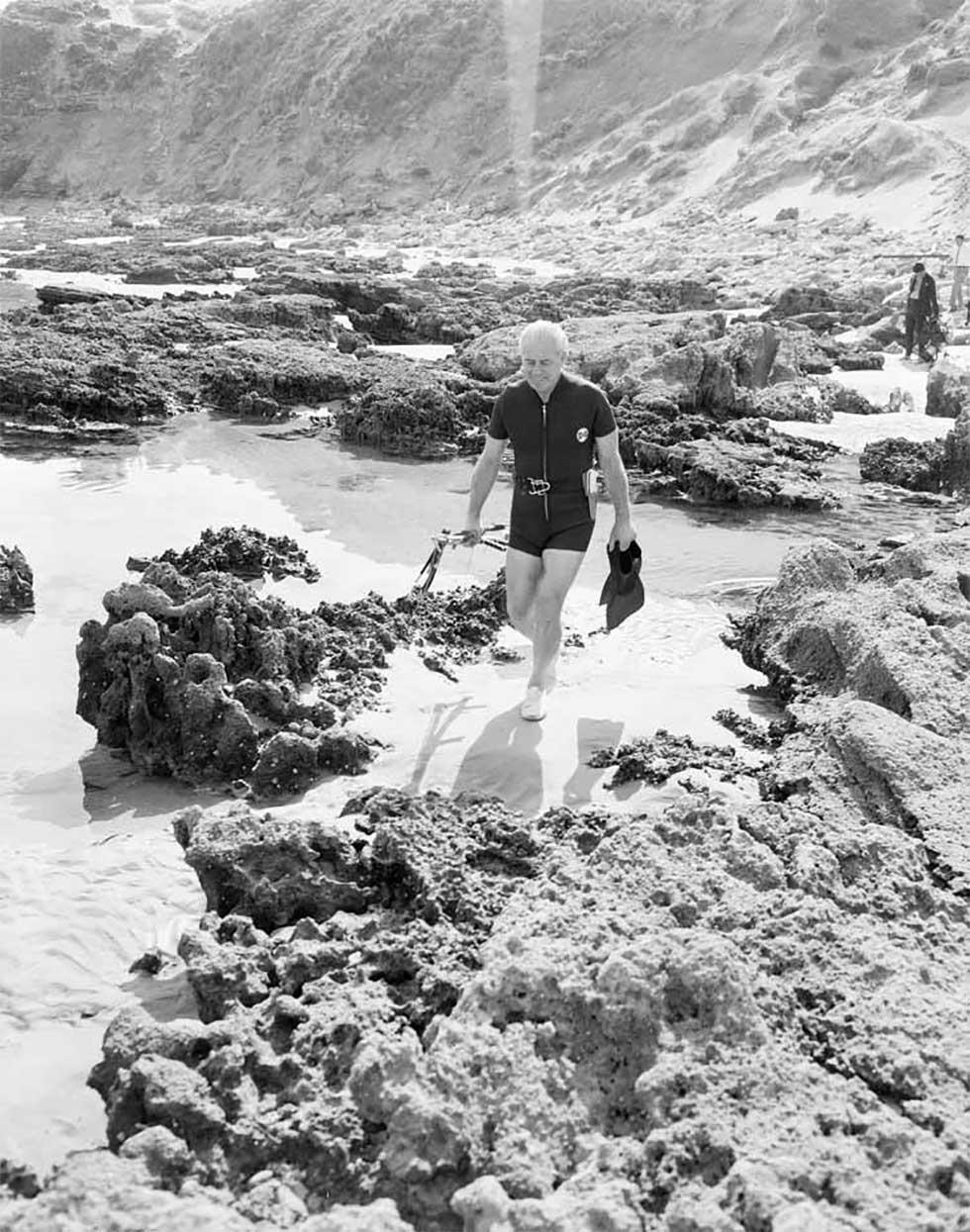 Harold Holt walking by rock pools wearing a spring wet suit, carrying flippers, snorkelling gear and a speargun.