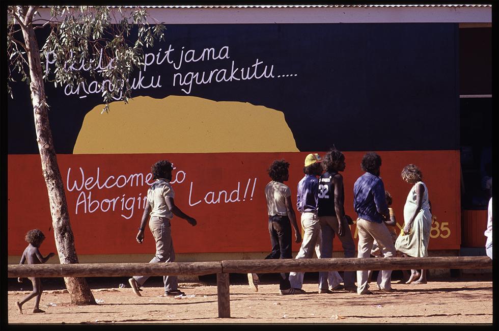 First Australians walk past a mural of the Aboriginal flag with the sun replaced by a yellow image of Uluru.