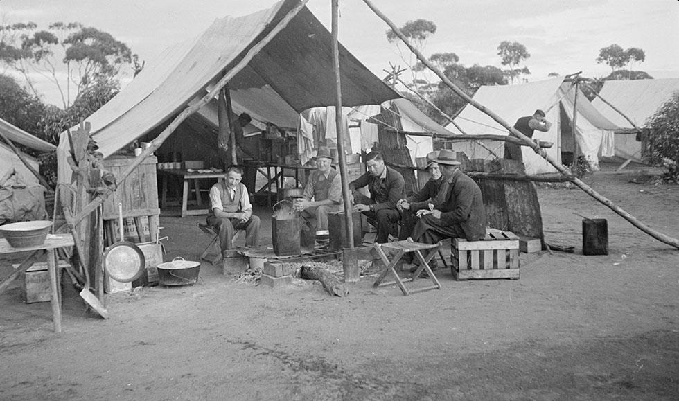 5 men sitting around a fire heating tin buckets in front of an open mess tent.