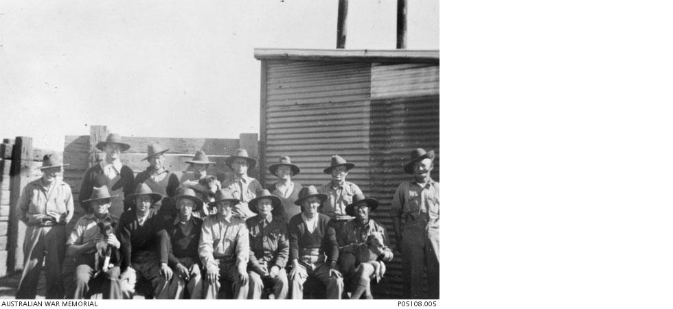 Assembled for a photo: 15 men and 3 dogs beside a tin shed and timber fence.