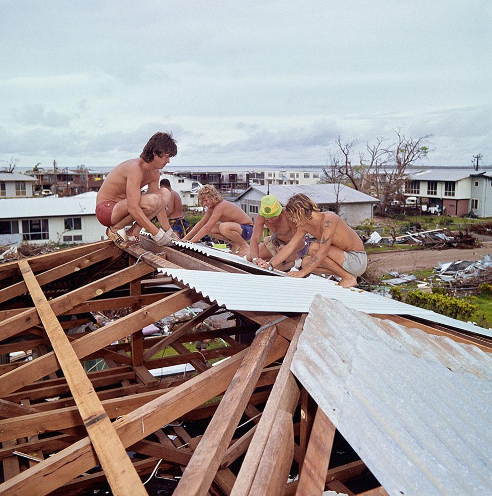 5 shirtless men in shorts nailing corrugated steel sheets onto battens.