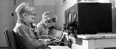 Two young boys at a lonely homestead do their lessons by transceiver radio during a School of the Air broadcast from Broken Hill, 1954.