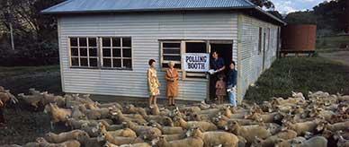 Five people standing near the doorway of a timber hall with a flock of sheep and lambs in the foreground.