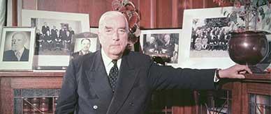 Robert Menzies standing in a wood-panelled room in front of a leadlight glass-fronted bookcase.