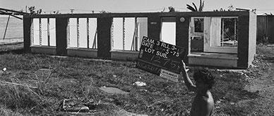 A man holding a house allotment sign infront of a damaged home.