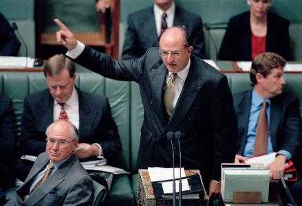 Peter Reith standing to addresses Parliament. Prime Minister John Howard seated and looking on.