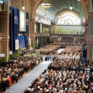 A mass of people seated inside the Royal Exhibition Building, Melbourne. Vaulted ceiling and large fanlight window above.