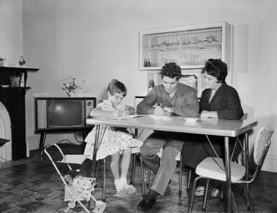 A man and his daughter write at a table, watched by his wife.