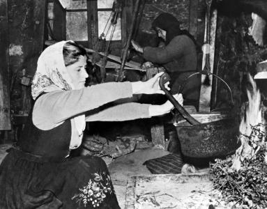 Vassiliski preparing food in a pot hung over a fire. A woman weaves in the background. 