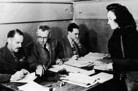 A woman stands in front of a seated selection committee of 3 men in suits. 