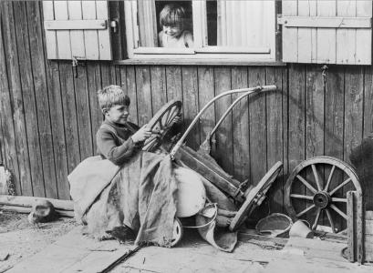 A young boy 'driving' a car made out of furniture and a pram.