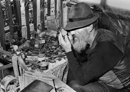 A man with a long beard and a fedora hat examines a watch at a table covered with watch pieces.