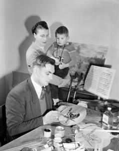 Otto painting a plate at his desk, with his wife watching holding their son on her hip.