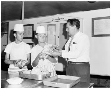 Laszlo Makay speaks to two cooks, in uniform, who are holding raw chickens. 