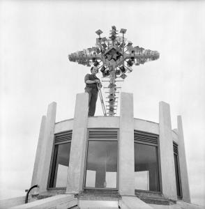 Voitre standing on the roof of a church next to an abstract cross made of welded metal. 