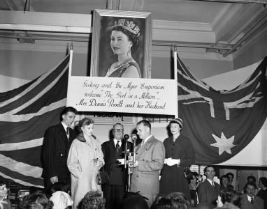 Barbara and Dennis Porritt being introduced on a podium. A portait of Queen Elizabeth II and a banner are above them.