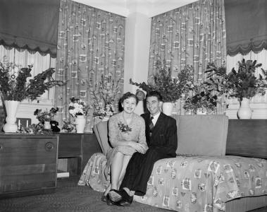 Barbara and Dennis portrait sitting on a floral couch, surrounded by vases of flowers.