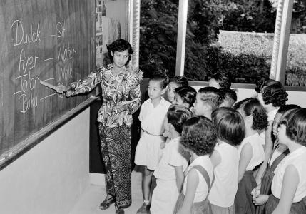 Ummi pointing at words on a blackboard observed by school children.