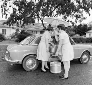 2 women, one holding an umbrella, place prepared meals into an Austin 1800 car.