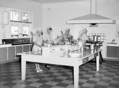 4 women prepare meals around a large table in a kitchen.