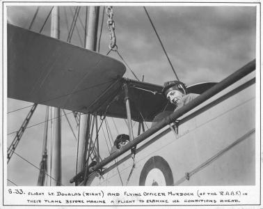 2 men looking down from the cockpit of a biplane aboard a ship.