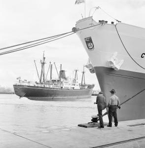 2 men stand beside the bough of a docked ship. While another ship moves past.